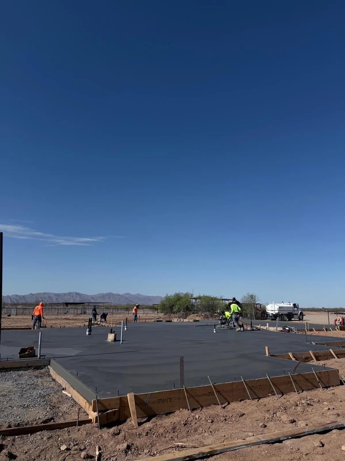 Crew finishing a large concrete foundation slab on an open lot, with wood forms around the edge and Arizona mountains on the horizon